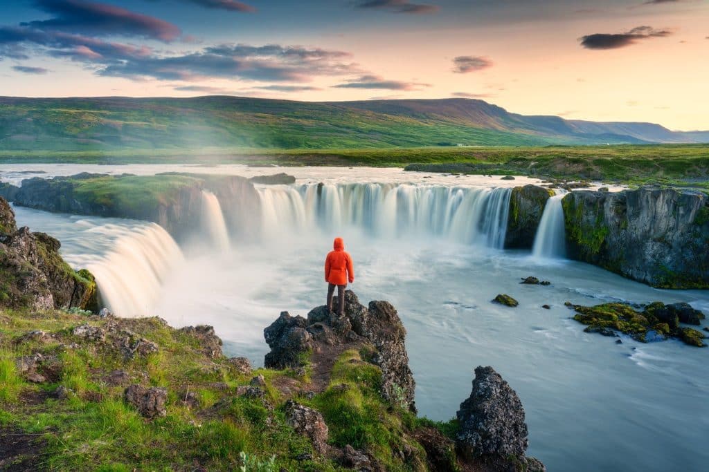 raveler in bright orange jacket overlooking Iceland's majestic Goðafoss waterfall at sunrise, capturing the raw beauty and peaceful isolation of a South Coast Iceland itinerary.