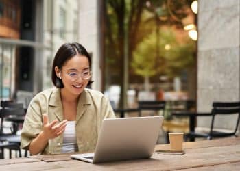 Smiling woman working remotely at an outdoor café with her laptop, representing modern places to work remotely near me with a relaxed atmosphere.