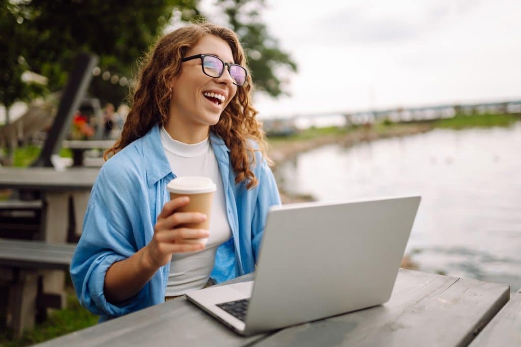 Happy freelancer working outdoors with a coffee, laptop, and a bright smile, enjoying the freedom of the daily routine freelancer lifestyle.