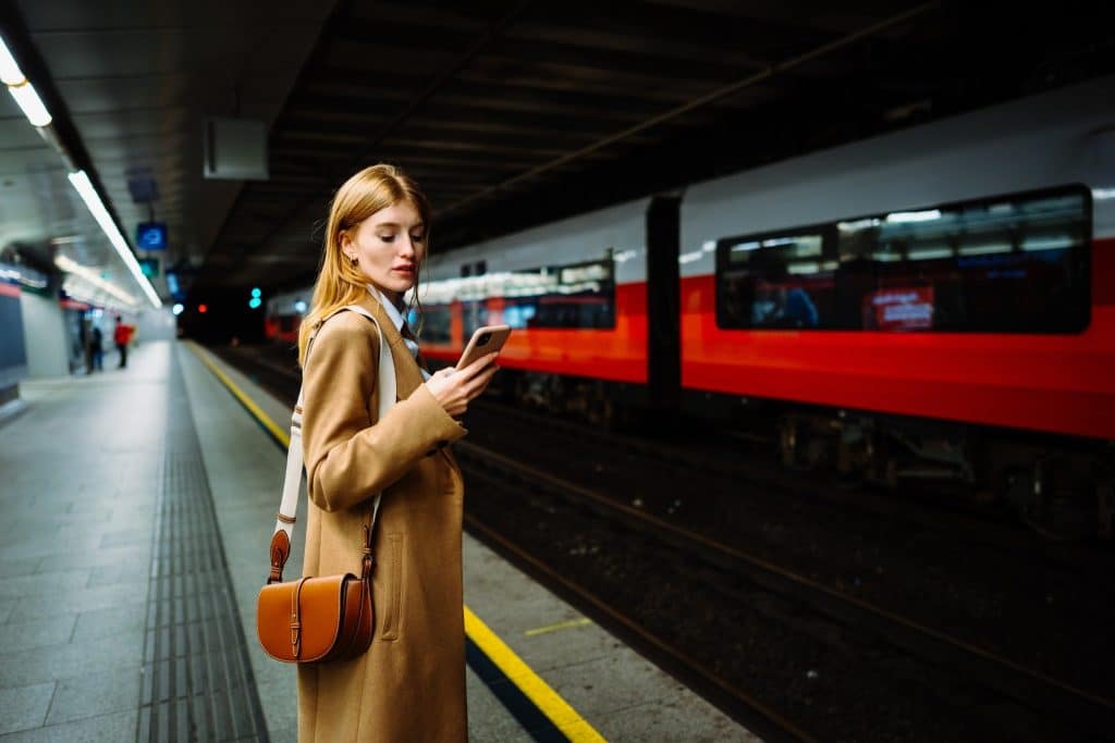 Young woman standing on a European train platform at night, checking her phone while waiting for a night train Europe departure — stylish and modern travel scene