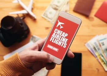 Hand holding a smartphone with a cheap airline flights booking app, surrounded by travel essentials and currency, representing smart tips for cheap flights.