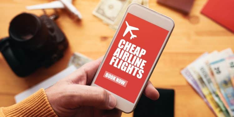 Hand holding a smartphone with a cheap airline flights booking app, surrounded by travel essentials and currency, representing smart tips for cheap flights.