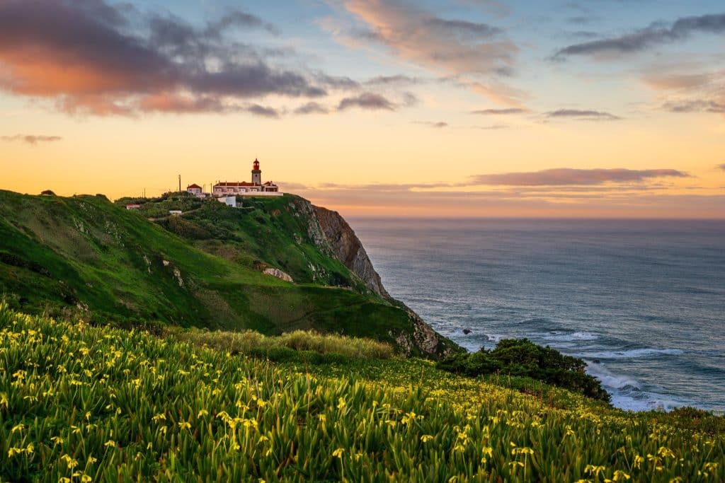 Stunning sunset view of Cabo da Roca lighthouse near Sintra, with dramatic cliffs, wildflowers, and the Atlantic Ocean, highlighting the beauty of one of the best cities in Portugal.