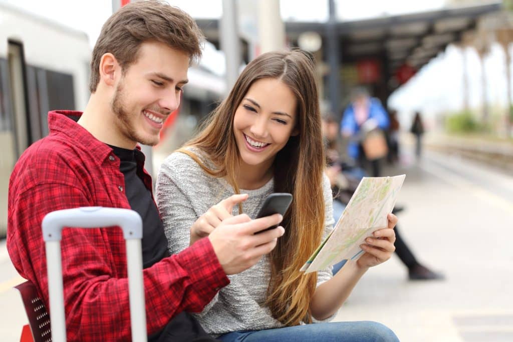 Smiling couple at a train station using a GPS and map potentially taking public transport to avoid common tourist traps.