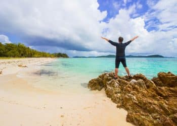 Traveler celebrating on rocky shore beside turquoise water and white sand dry-season vibes from the best Philippine islands.