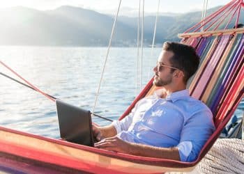 A digital nomad relaxing in a hammock by the sea while working on a laptop, showing how digital nomad productivity tips help balance work and travel.