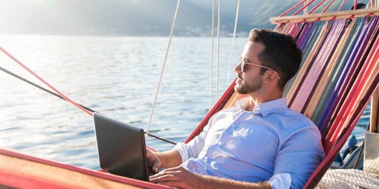 A digital nomad relaxing in a hammock by the sea while working on a laptop, showing how digital nomad productivity tips help balance work and travel.