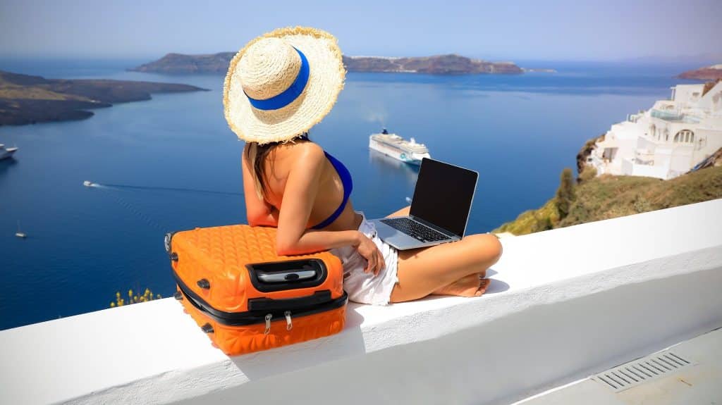 A happy digital nomad sitting with a laptop and suitcase overlooking the sea, relaxing and capturing digital nomad productivity tips for combining work with travel freedom.