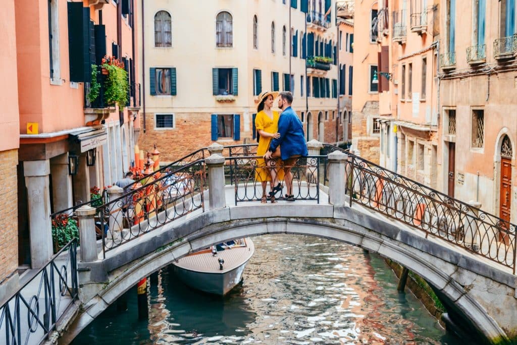 A couple standing on a small bridge over a canal surrounded by colorful Venetian buildings.