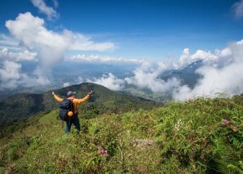 Solo traveler with backpack raising arms on a mountain ridge, symbolizing the freedom of exploring the world on a solo travel budget.