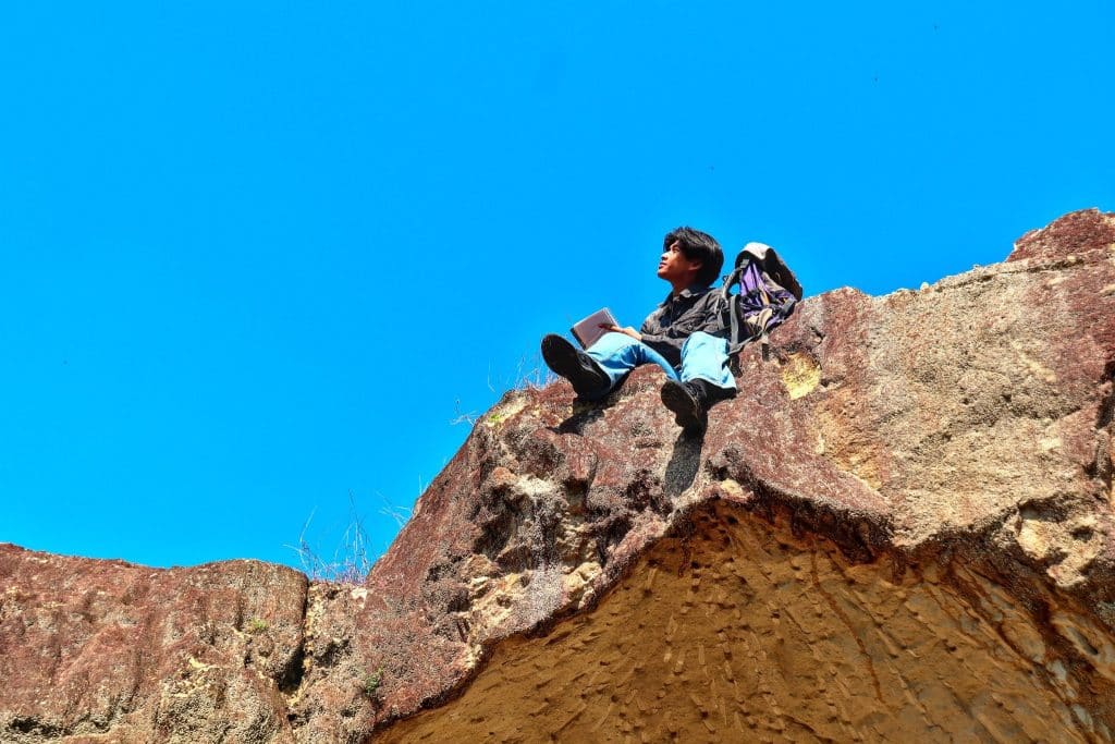 Traveler sitting on a rocky cliff with backpack and notebook, reflecting on experiences made possible by a well-planned solo travel budget.